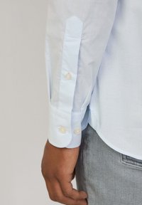 Light blue checkered shirt featuring a buttoned cuff with two round, white buttons and a rounded hemline. The fabric appears smooth and lightweight.
