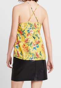 Camisole florale jaune à bretelles croisées dans le dos, ornée de fleurs roses et bleues vibrantes. Associée à une mini-jupe en cuir noire.