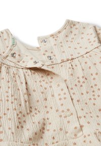 Bežový látkový oděv s texturovaným povrchem, který má světle hnědé tečkované vzory, cvoky na límci a nařasené detaily.