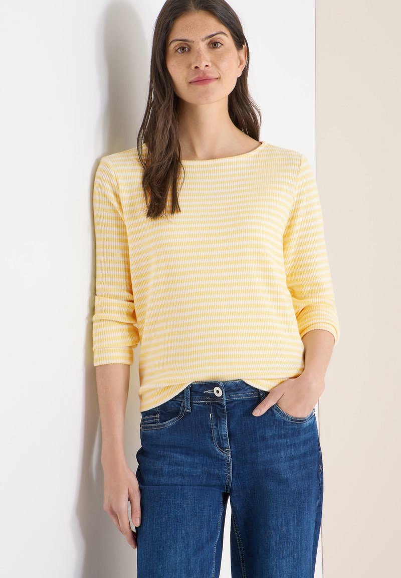 Haut à manches longues rayé jaune et blanc avec un col bateau, associé à un jean en denim bleu foncé. Texture lisse, coupe décontractée.