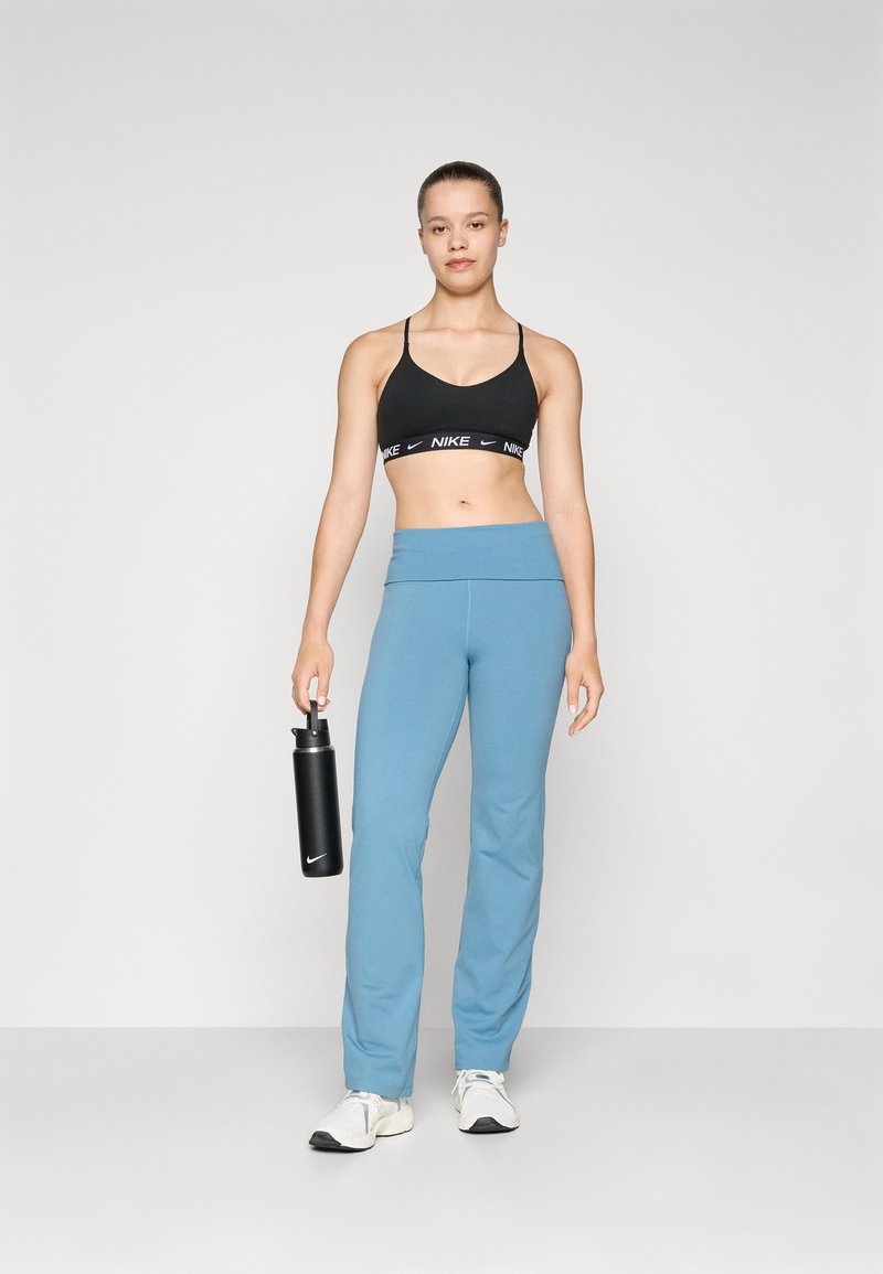 Reggiseno sportivo nero con logo, pantaloni flessibili azzurri, tiene in mano una bottiglia d'acqua nera e indossa sneakers bianche su uno sfondo neutro.