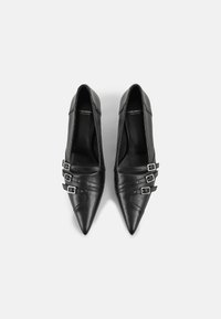 Chaussures en cuir noir à bout pointu avec trois brides argentées bouclées sur le devant, vues de dessus sur un fond gris clair.
