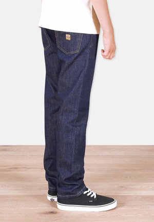 Person wearing dark blue straight-leg jeans and black low-top sneakers, standing on light wood floor against white background.