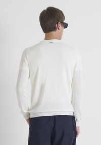 Maglione bianco a maglia con accenti a coste sulle spalle, scollatura rotonda e maniche lunghe. Indossato con pantaloni scuri, visto da dietro.