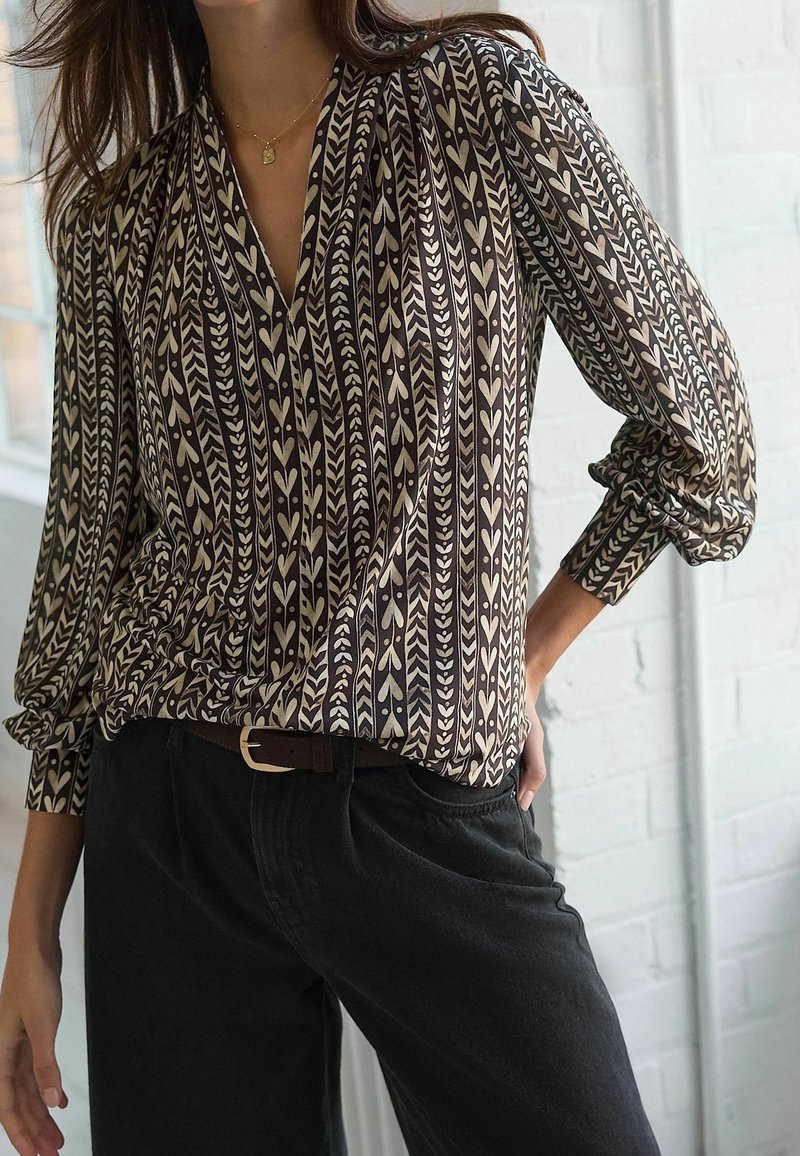 Femme portant un chemisier à manches longues à motifs beige et noirs avec des motifs verticaux de cœurs et de feuilles, associé à un pantalon noir et une ceinture marron.