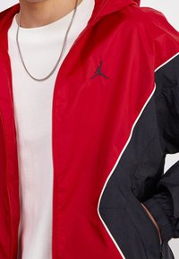 Červeno-černá větrovka s kapucí, vybavená předním zipem, bílým zdobením a černým logem na hrudi.