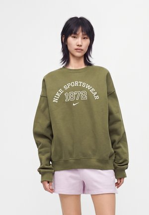 Ung person med sort hår iført olivengrøn Nike Sportswear sweatshirt og lyse lilla shorts, stående mod en ensfarvet baggrund.