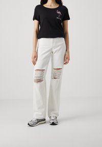 Kvinna i svart T-shirt med rosa flamingodesign och vita rivna jeans, matchat med silvriga och svarta sneakers, står på vit golv.