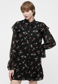 Νεαρή γυναίκα με κοντά μαλλιά, φορώντας μαύρο φλοράλ φόρεμα με μακριά μανίκια, ρίγες και κουμπιά, πόζαρε με το ένα χέρι στη μέση της μπροστά σε λευκό φόντο.