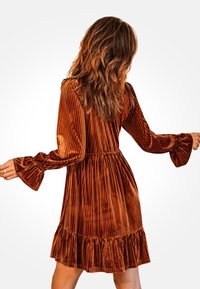 Vestido de terciopelo color óxido con rayas verticales, mangas largas acampanadas y dobladillo con volantes. Cintura ajustada, textura suave y diseño elegante.