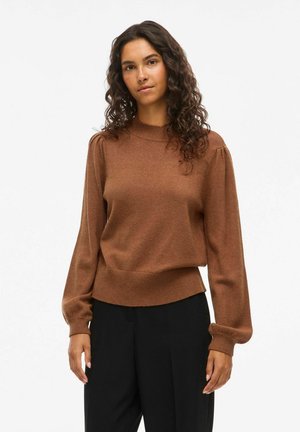 Femme aux cheveux bouclés et foncés, portant un pull marron à manches longues et un pantalon noir, debout devant un fond blanc uni.