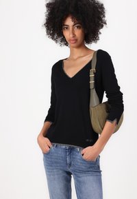 Maglione nero con scollo a V e rifinitura argentata sul colletto e sui polsini, abbinato a jeans in denim azzurro. Il modello tiene una borsa verde a tracolla.