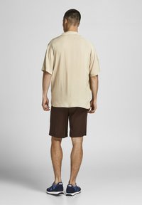 Camisa beige de manga corta y pantalones cortos marrones, con un corte relajado. Los zapatos son navy con acentos azules, se muestra la vista trasera sobre un fondo claro.