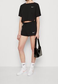 Woman wearing black Nike cropped T-shirt and shorts, white Nike socks and sneakers, holding a shiny black handbag against a white background.