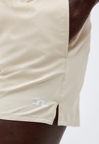 Μπεζ σορτς με πλαϊνή σχισμή και μικρό λευκό λογότυπο, φορεμένα σε σκουρόχρωμο δέρμα, που δείχνουν την ομαλή υφή του υφάσματος και τις λεπτομέρειες της ραφής.