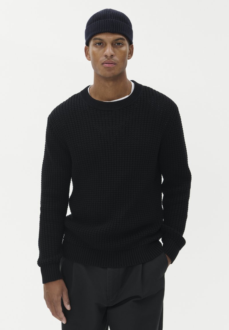 Man som bär en svart stickad tröja, svart stickad mössa och svarta byxor, står med en hand i fickan framför en enkel vit bakgrund.