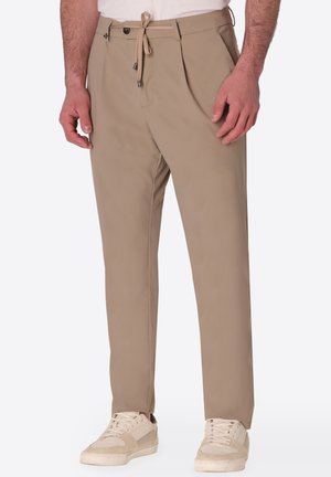 Uomo che indossa pantaloni beige a taglio stretto con pieghe, coulisse, passanti per cintura e scarpe da ginnastica bianche.
