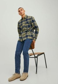Camisa de franela a cuadros en verde y azul, con dos bolsillos en el pecho; vaqueros ajustados; zapatos de ante color marrón; sentado en una silla de madera con patas de metal negro.