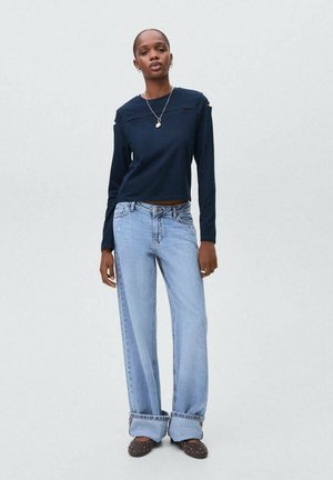 Frau in einem marineblauen Langarmoberteil, hellblauen, weit geschnittenen Jeans mit Umschlägen, einer Halskette und schwarzen, mit Nieten besetzten Slippern, die vor einem einfarbigen Hintergrund steht.