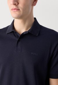 Tamsiai mėlyna polo marškinėliai, pagaminti iš minkštos medžiagos, su dviem mygtukais, apykakle ir diskretišku „BOSS“ logotipu, išsiuvinėtu toniniu siūlu.