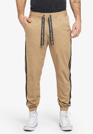 Pantalones joggers beige con franjas negras laterales y cordones negros con texto blanco, combinados con zapatillas deportivas altas blancas.