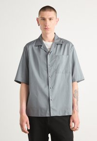 Camisa de manga corta, con botones, en tela gris sólida que presenta un bolsillo de parche, cuello clásico y textura suave.