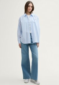 Camisa azul claro de gran tamaño con cuello puntiagudo, combinada con jeans de pierna ancha. Textura suave, diseño clásico y ajuste casual.