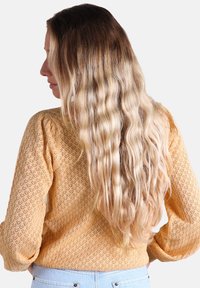 Pull en tricot jaune avec un motif en losange texturé, cheveux blonds ondulés et longs tombant sur l'épaule, jeans bleu clair.