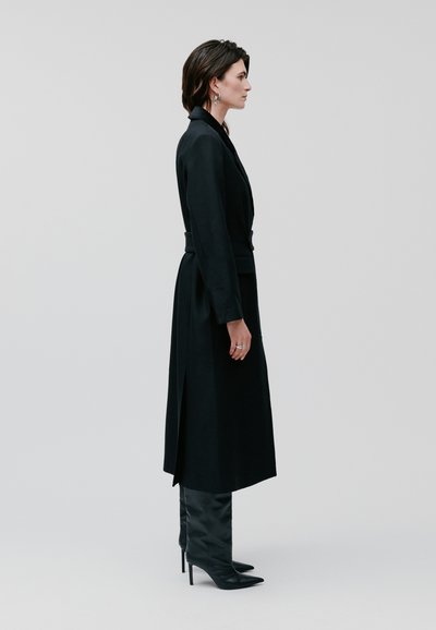 Cappotto nero su misura con maniche lunghe e uno spacco laterale, abbinato a stivali neri con tacco a spillo fino al ginocchio. Tessuto liscio, design aderente.