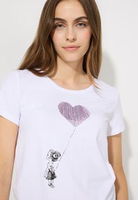 Weißes T-Shirt mit einer schwarz bedruckten Illustration eines Mädchens, das einen rosa herzförmigen Ballon hält, mit kurzen Ärmeln und Rundhalsausschnitt.