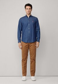 Donkerblauwe denim overhemd met een kraag, lange mouwen en een klein geborduurd logo op de borst; gecombineerd met tan chinos.