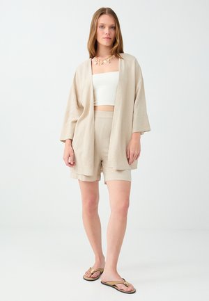 Kobieta ubrana w beżową kurtkę w stylu kimono, pasujące szorty, biały top bandeau, złoty naszyjnik i beżowe klapki, stojąca na białym tle.