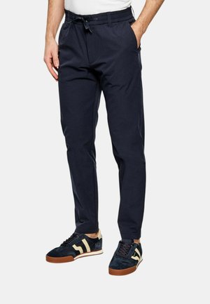 Man wearing navy blue casual pants with drawstring waist and navy sneakers with beige accents, standing with hands in pockets.