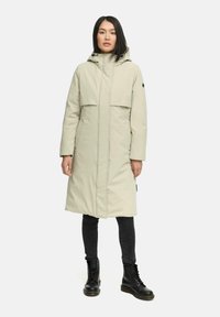 Lang lichtbeige buitenkleding met een capuchon, voorkruiszakken en een rechte snit. Draag het over zwarte jeans en zwarte veterschoenen.
