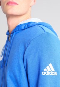 Sweat à capuche bleu en tissu doux, doté d'une fermeture zippée, d'une capuche doublée, et d'un logo Adidas blanc sur la manche gauche.