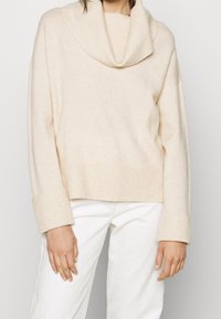 Beige ribbstickad tröja med trekantskrage och breda muddar, kombinerad med vita byxor, beskuren bild med fokus på bålen och händerna.