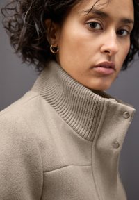 Beżowa kurtka z mieszanki wełny z wysokim żeberkowanym kołnierzem, zapięciem na napy i strukturalnym wykończeniem, zaprojektowana z myślą o cieple i komforcie.