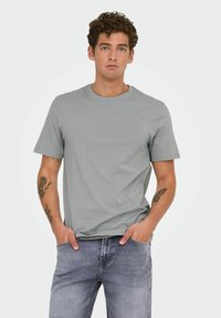 Camiseta de algodón gris con escote redondo y mangas cortas, combinada con jeans de mezclilla claros. El fondo es blanco liso.