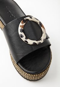 Sandale glissante en cuir noir avec un accent rond crème décoratif et à motif. Semelle épaisse en jute tissée qui ajoute de la hauteur et de la texture.