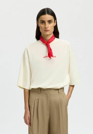 Femme aux longs cheveux foncés portant une chemise blanche oversize, un pantalon beige plissé et un foulard rouge autour du cou, debout devant un fond uni.