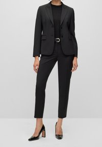 Černý dokonalý blazer s klopami, spárovaný s úzkými černými kalhotami a lodičkami s ostrou špičkou. Hladká látka s minimálním zdobením.
