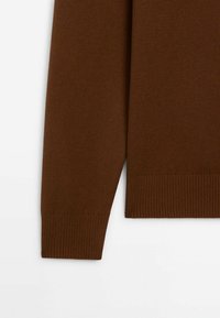 Manche et ourlet d'un pull marron à manches longues avec poignets et bord inférieur côtelés, présenté sur fond blanc.