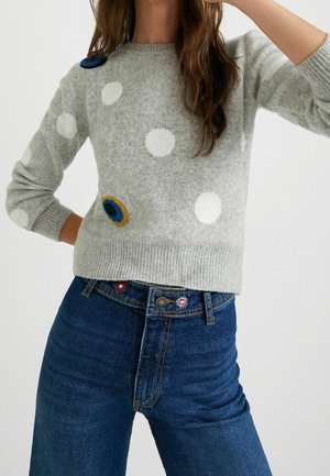 Grijze sweater met witte stippen en levendige geborduurde accenten. Geribbelde manchetten. Combinatie met high-waisted blauwe jeans met bloemgeborduurde details.