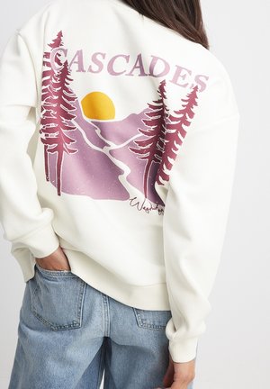 Personne portant un sweat-shirt blanc avec un graphisme de montagne violette, de pin rouge et de soleil jaune, ainsi qu'un texte indiquant Cascades et Washington.