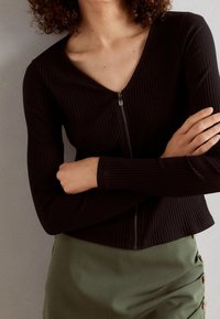 Top a maniche lunghe nero a costine con scollatura a V e dettaglio con zip, abbinato a una gonna verde con bottoni laterali.