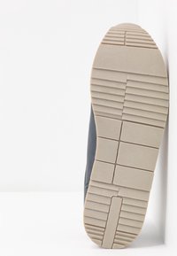 Semelle d'une chaussure beige et grise avec un profil antidérapant à motif, appuyée contre un mur blanc.