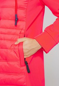 Red padded jacket featuring a smooth fabric finish and a hidden side pocket with a contrasting zip. Drawstring hood with adjustable toggle.