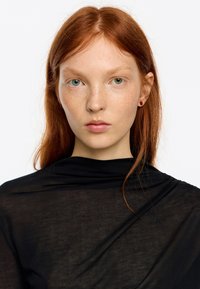 Modelka z długimi rudymi włosami ubrana w czarną, przezroczystą bluzkę. Na twarzy widoczne charakterystyczne piegi, zielone oczy oraz małe, czerwone kolczyki.