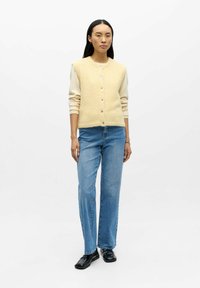 Kremfarget ullblandet ermeløs cardigan med knappelukking, brukt over en lys genser og kombinert med lyseblå jeans med rette ben og svarte loafers.