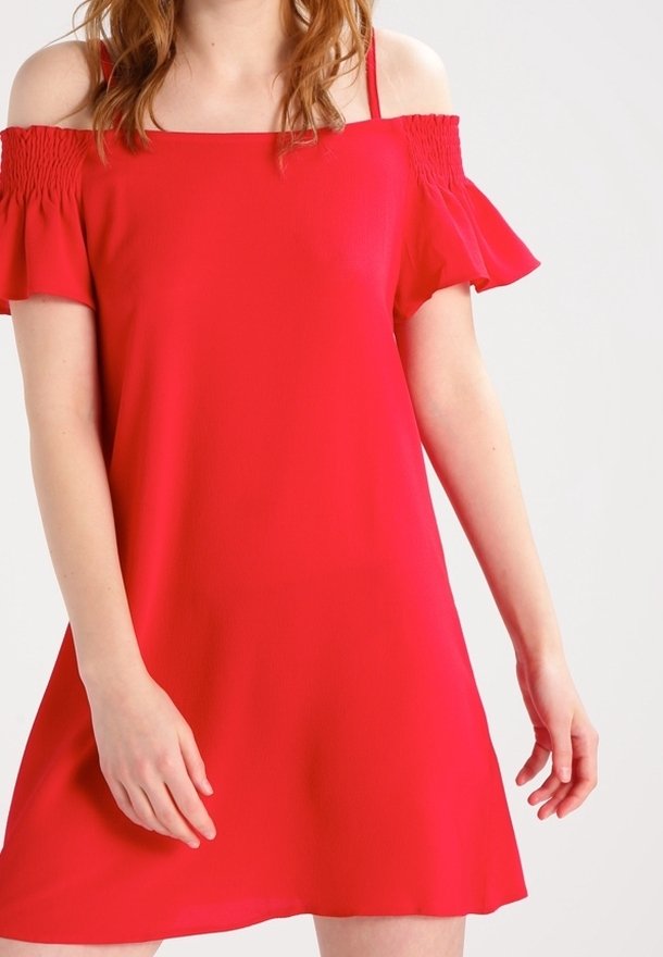 Femme portant une robe rouge vif épaules dénudées avec des manches courtes à volants sur un fond clair uni.